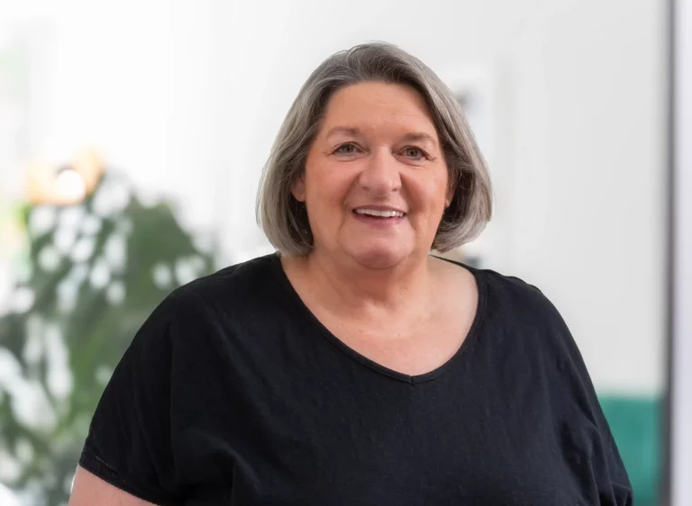 Lächelnde Frau mit grauen Haaren, die ein schwarzes T-Shirt trägt, in einem hellen Raum.