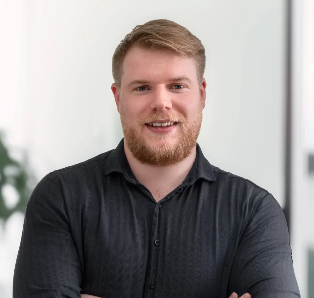 Mann mit blonden Haaren und Bart, der in die Kamera lächelt und ein schwarzes Hemd trägt.