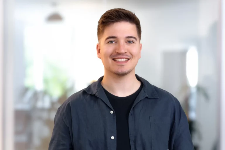 Junger Mann mit kurzem, braunem Haar und einem freundlichen Lächeln, gekleidet in einem schwarzen T-Shirt und einer blauen Jacke.