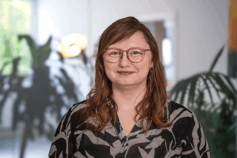 Portrait einer Person mit Brille und braunen Haaren, die in einem Raum mit Pflanzen steht.