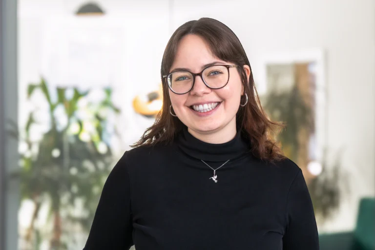 Lächelnde Frau mit Brille und schwarzem Oberteil, die in einem modernen Büro steht.