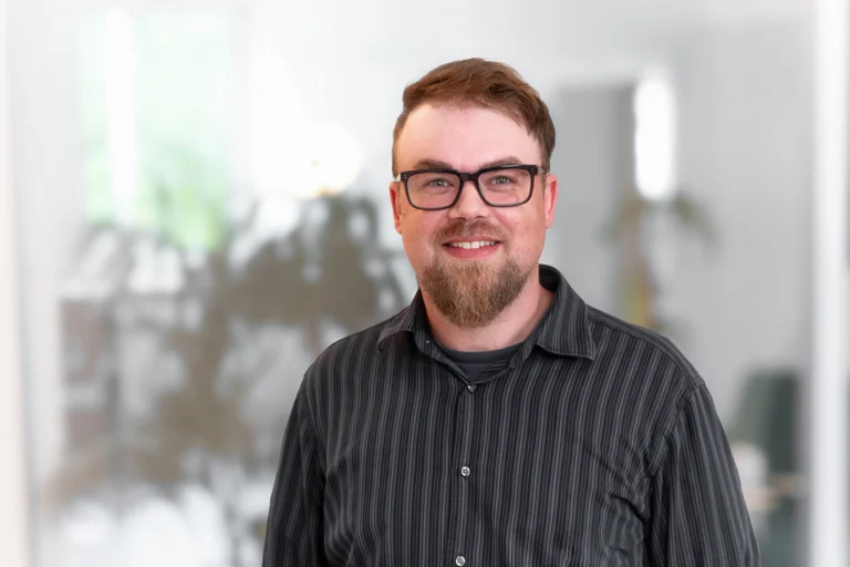 Portrait eines Mannes mit dunkelgrau gestreiftem Hemd und Brille, braune Haare und Bart, in einem modernen Büro hinter Glaswand, Name Alex