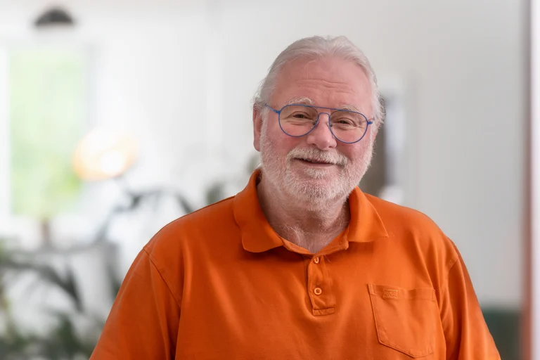 Dieter im hellen Büro, orangefarbenes Polohemd, blaue Brille, graue Haare und freundliches Lächeln; unscharfer Hintergrund
