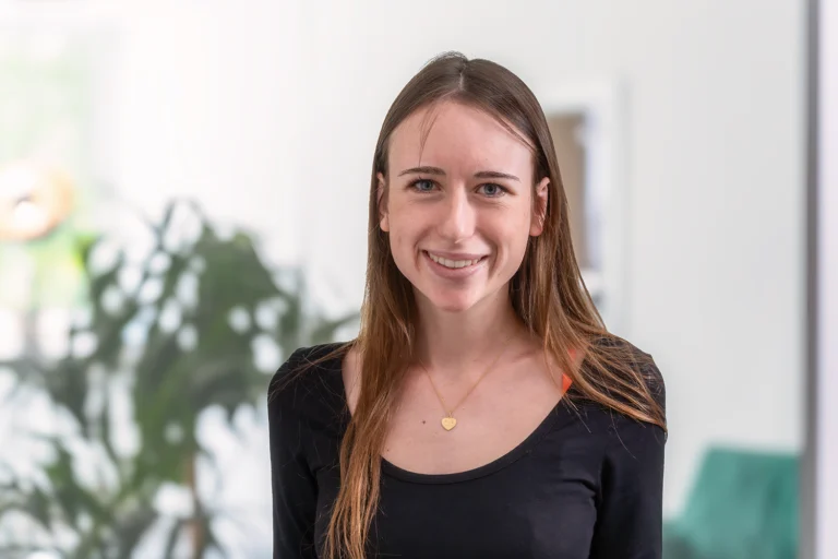 Porträt von Nadine, einer jungen Frau mit braunen Haaren, schwarzem Oberteil und goldenem Herzanhänger, vor hellem Bürohintergrund mit dezentem Grün im Hintergrund.