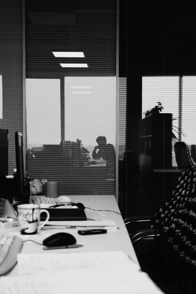 Schwarz-weiß Büroaufnahme mit Blendenfenstern, Schreibtisch, Maus, Tasse und Unterlagen; Silhouette zweier Personen im Hintergrund sowie Pflanzen, aufgenommen von Max Karpyza