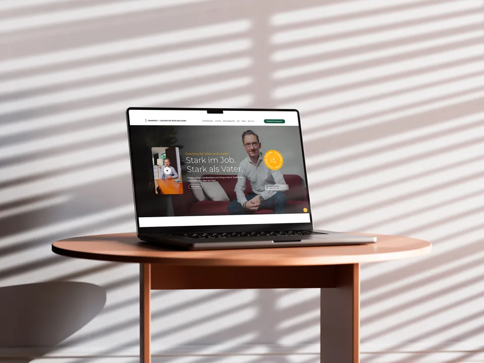 Laptop auf einem runden Holztisch vor einer Wand mit Streifenschatten, auf dem Bildschirm der Einstieg der DadMindset-Landingpage mit Videokarte und Porträt des Coaches.